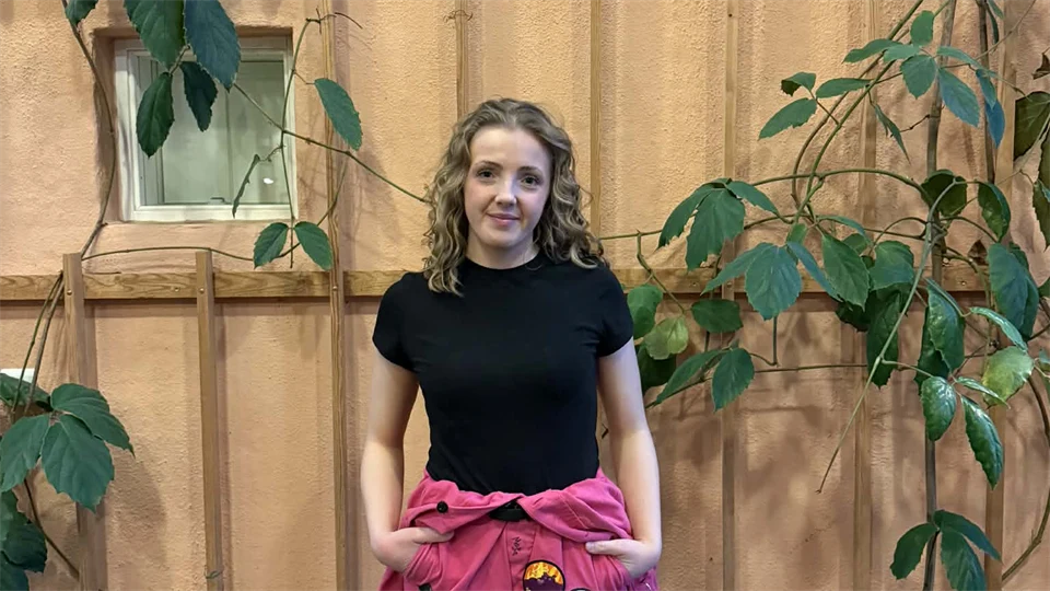 Ung kvinna med svart kortärmad tröja och ceriserosa overall knuten runt midjan står framför en beige vägg med gröna växter som slingrar sig. 