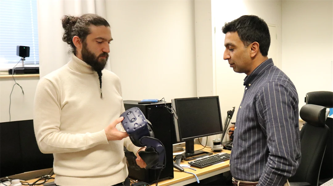  Two men stand together in a computer room. One man is holding a pair of VR glasses.
