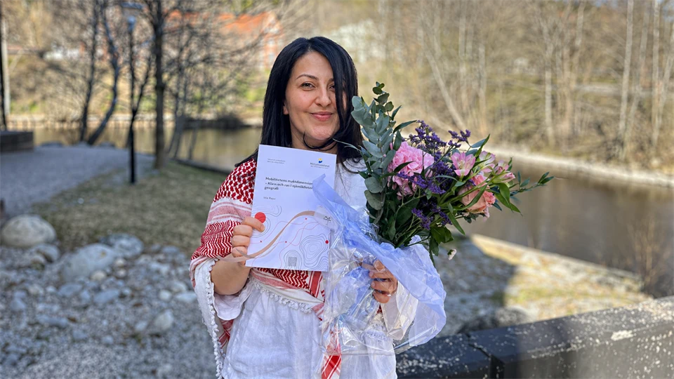 Kvinna står utomhus hållandes blommor och ett diplom.