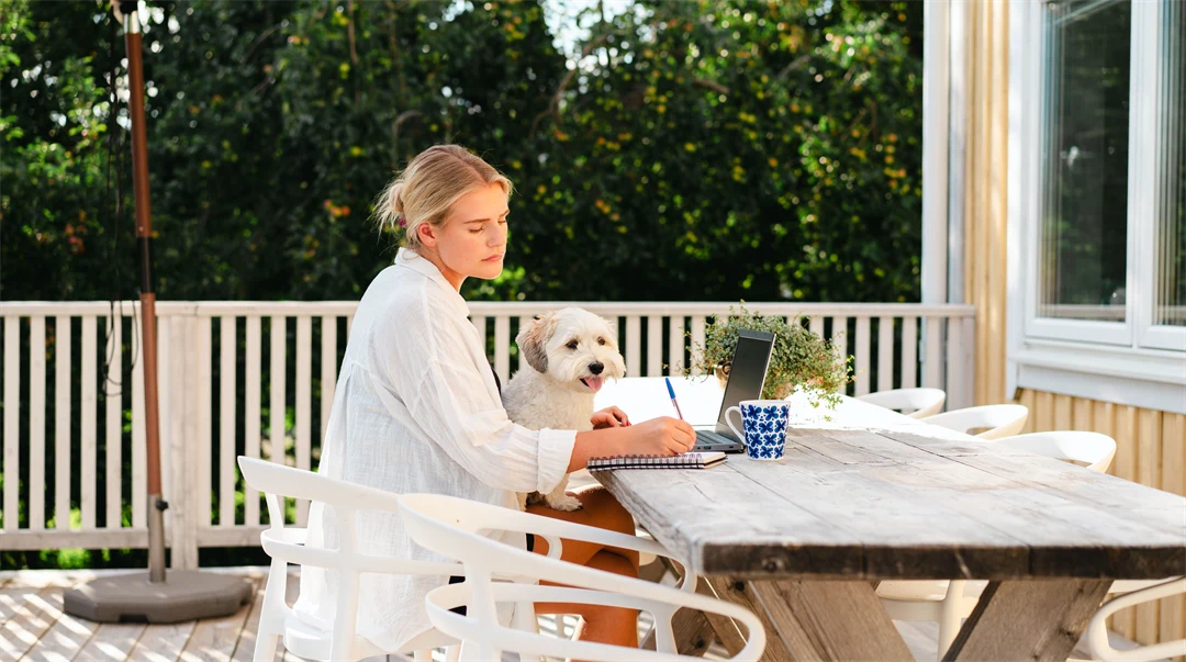 Kvinna som sitter vid ett bord på altan med laptop och pluggar. Har en hund i knät. 