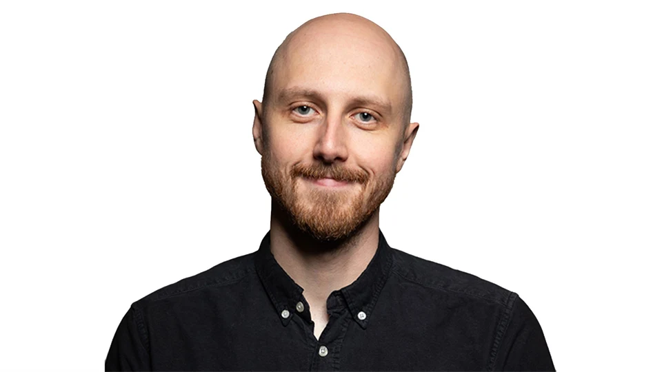 Man med skägg och mustasch ler mot kameran, klädd i svart skjorta, vitt bakgrund.