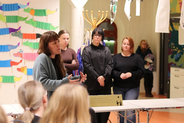 En grupp människor lyssnar på en kvinna som sitter på en bord längst fram i en sal. På väggen i bakgrunden syns färgglada alster skapade av barn. 