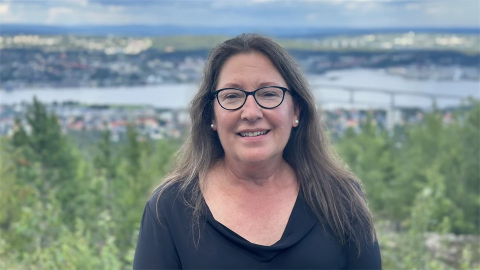 En mörkhårid kvinna med glasögon. Bakom henne ser man skog, byggnader, hav och en bro.