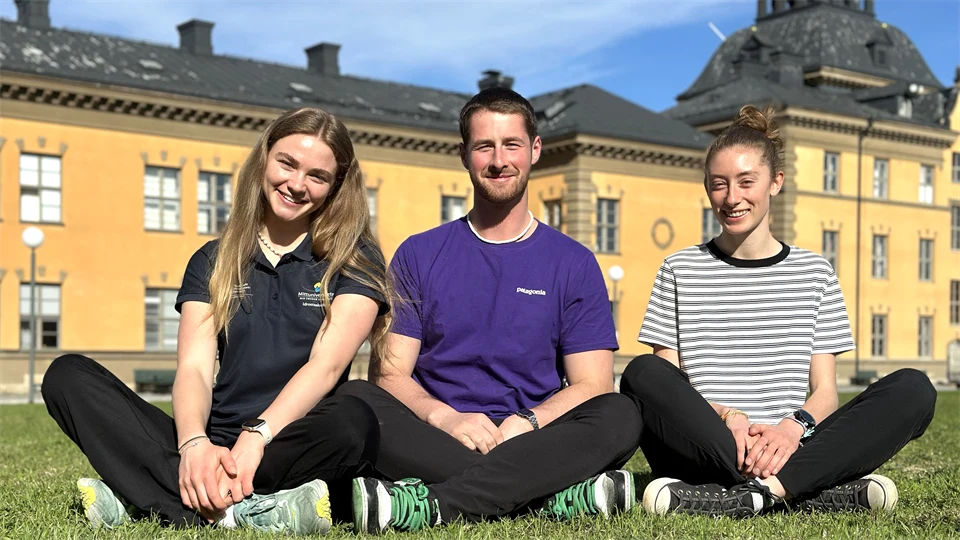 Tre unga människor sitter på gräs framför gul byggnad.
