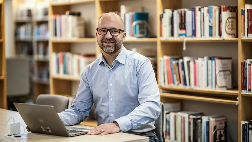 Man sitter vid ett bord i ett bibliotek. Han har en dator framför sig och bokhyllor bakom sig. Mannen bär en blå skjorta och är glad.