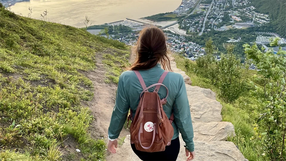En kvinna med ryggsäck på ryggen går på en stenbelagd stig och ser ut över en norsk fjord.