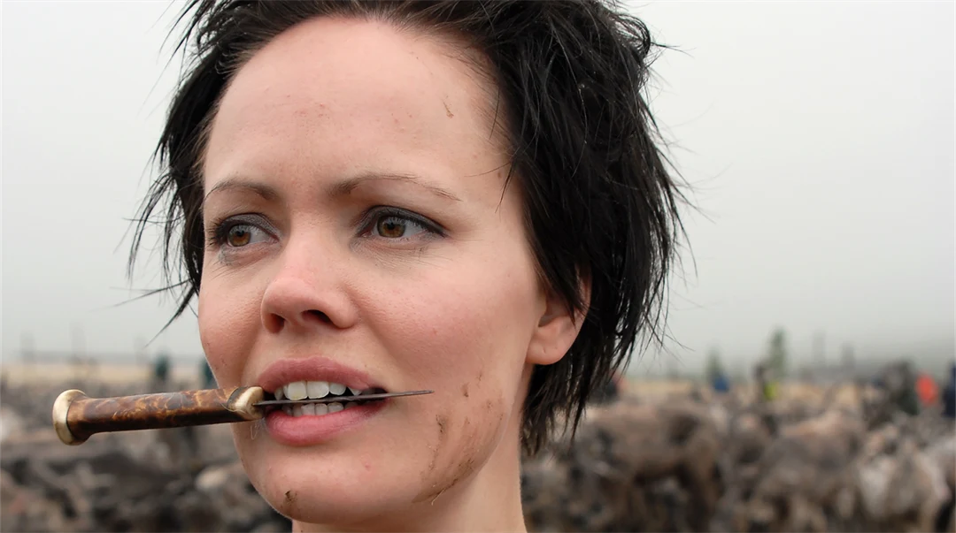 Kvinna i svart topp med spretig frisyr håller en kniv mellan tänderna.