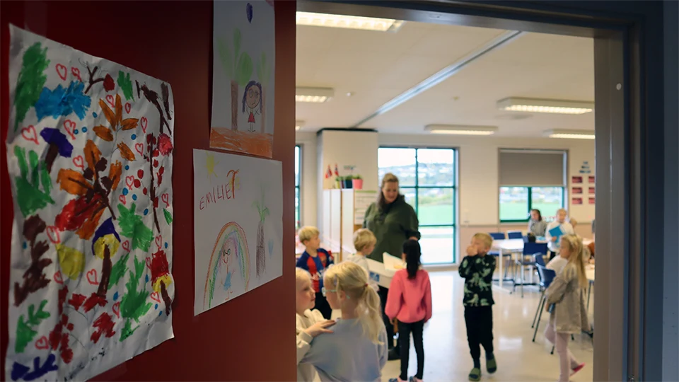En vy av ett klassrum genom en öppen dörr. Inne i klassrummet syns en kvinnlig lärare och omkring tio barn. I förgrunden till vänster syns en röd vägg med tre teckningar