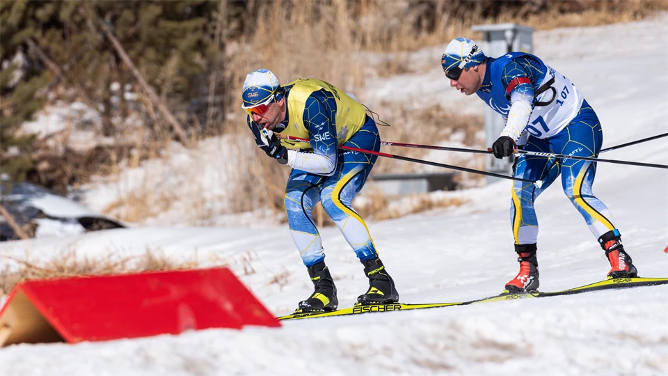 Två skidåkare i blågula dräkter som åker utför en bana märkt med röda v-boards. Den bakre skidåkaren håller i den främre åkarens ena skidstav.