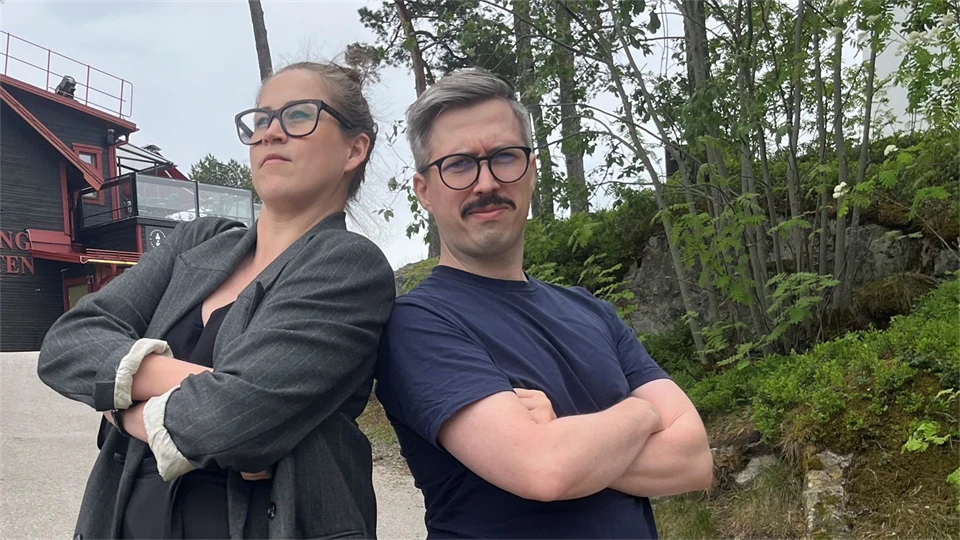 En kvinna och en man står bredvid varandra med lite träd och en byggnad i bakgrunden. 