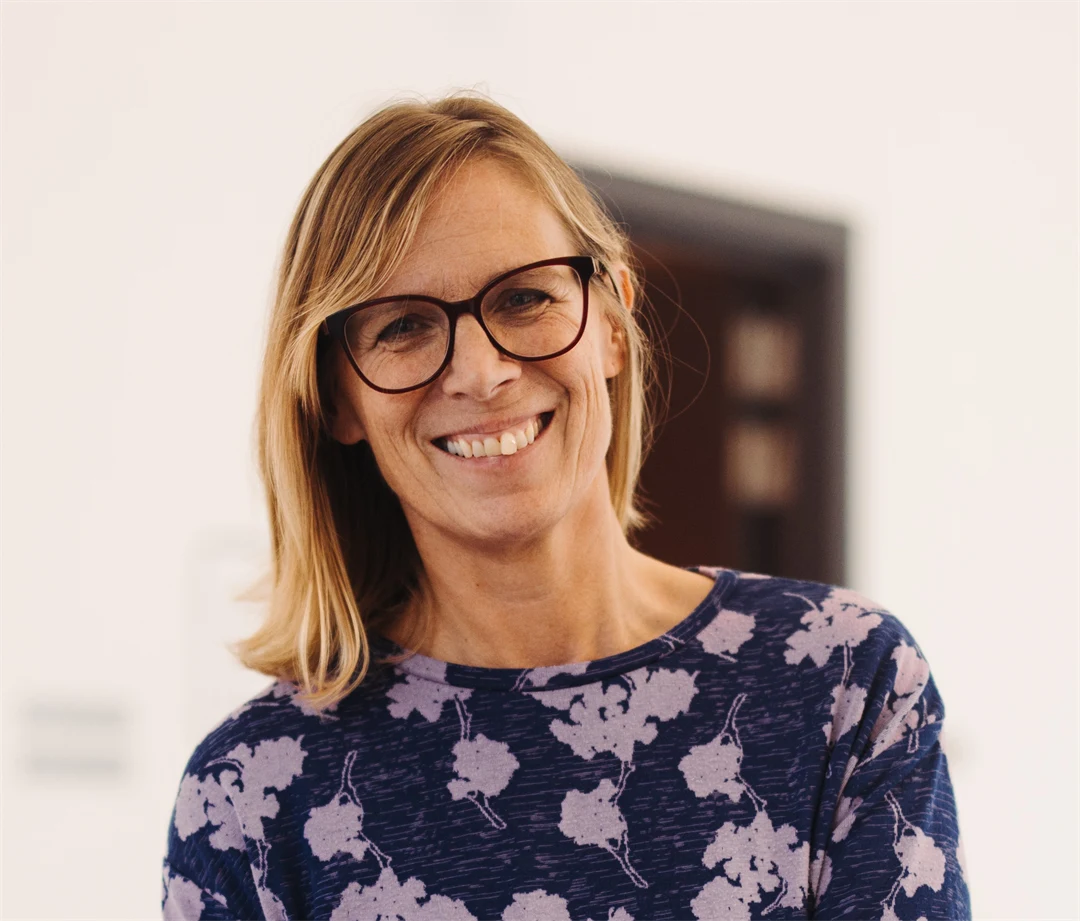 En person med axellångt blont hår står inomhus i en ljus miljö. Personen bär en mörkblå tröja med ett mönster av ljusa blommor.