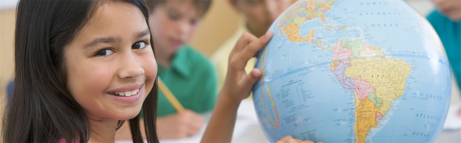 Fyra barn sitter vid ett bord. Tre av dem skriver och en av dem, en flicka med långt mörkt hår och rosa t-shirt pekar på jordglob.