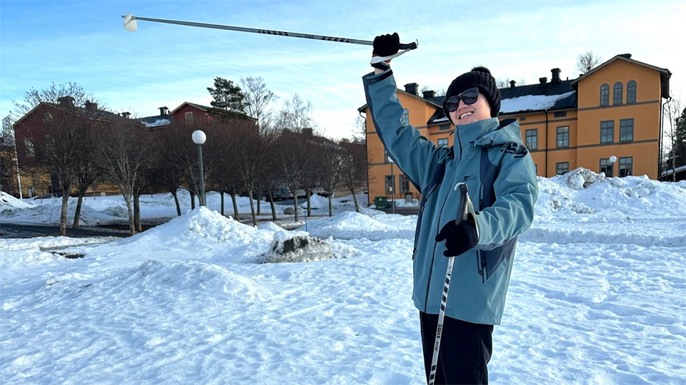 Young man with poles in his hands stands on a snow-covered field and raises one hand in the air and smiles