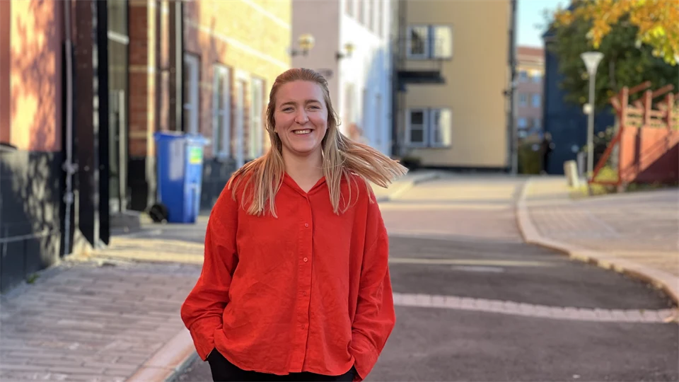 A woman in a red blouse looks into the camera and smiles. She has a street and some house facades in the background.