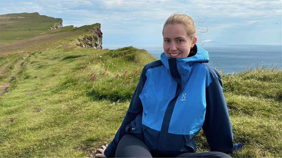 Ung kvinna i blå vindjacka, sittandes på en bergskam med havet nedanför. Hon är glad.
