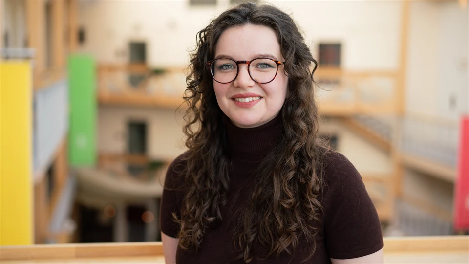 En kvinna med mörkt, långt, lockigt hår, glasögon och en mörkbrun kortärmad polotröja. 