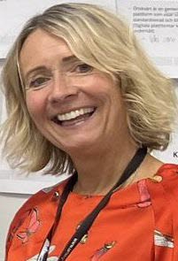 En kvinna i blont axellångt hår, svart nyckelband runt halsen och orange blus med fjärilar på. 