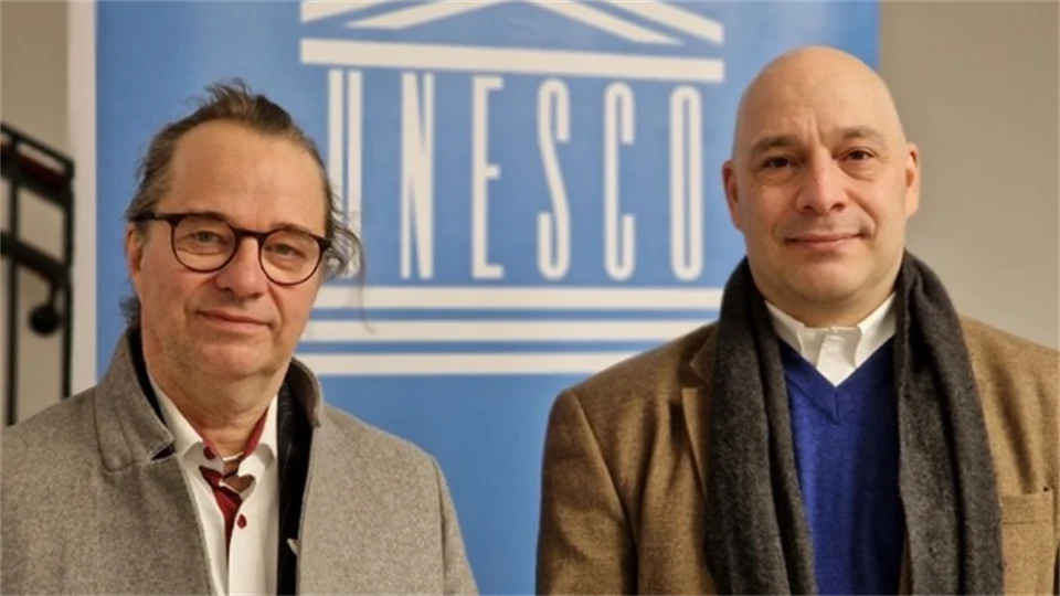 Två män klädda i rockar står framför en blå skylt med UNESCO-logotyp 