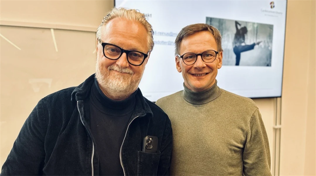 Två män i glasögon tittar in i kameran och ler. De står framför en skärm. 