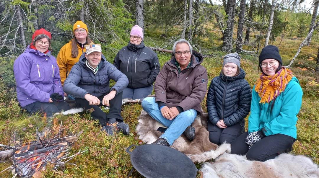 Sju personer sitter på renskinn invid en eld i skogen.