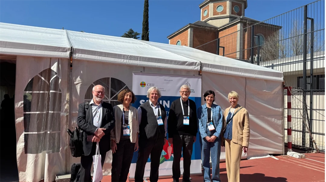 Six people dressed in costumes stand in front of a large white party tent. They have lanyards around their necks and in the background is a church-like building.