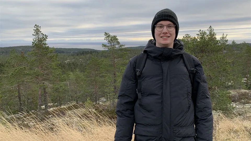 Ung kille i svart jacka och svart mössa som står framför små granar uppe på en höjd.