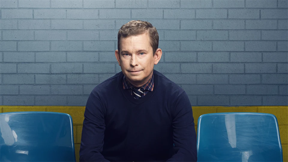 En man sitter på en stol i en stolsrad framför en blå och gul vägg.