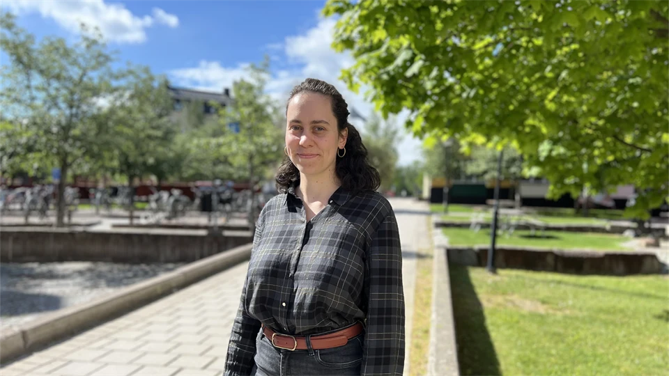 A woman is standing on a cobblestone path, looking directly at the camera. Trees can be seen in the background.