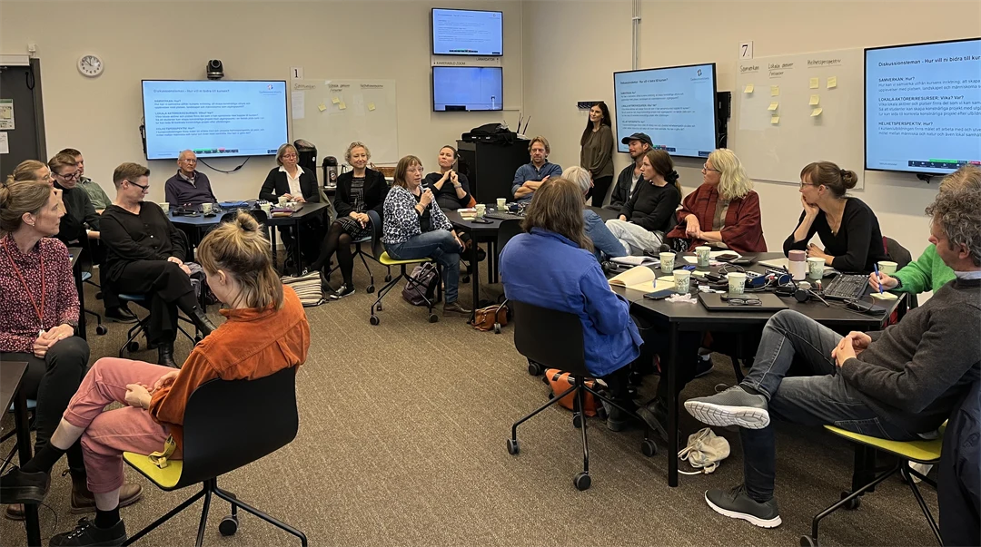 Ett trettiotal personer sitter vid runda bord i ett rum och pratar i mindre grupper. 