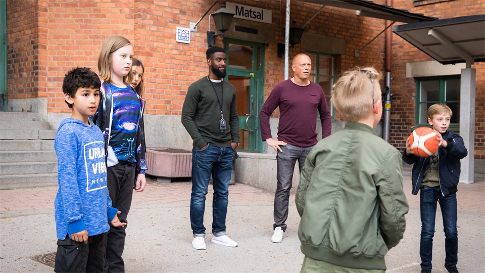 Barn leker med boll på skolgård, två vuxna närvarande.