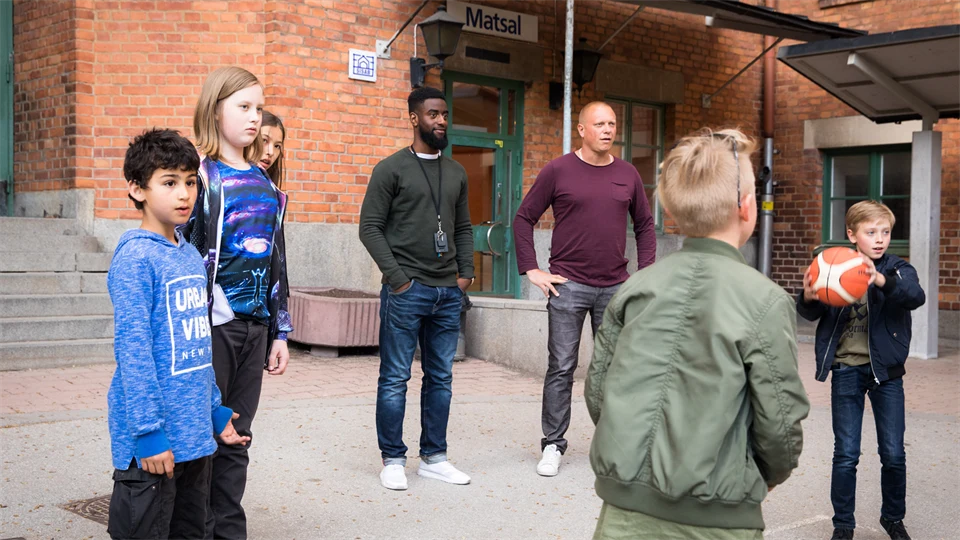 Barn leker med boll på skolgård, två vuxna närvarande.