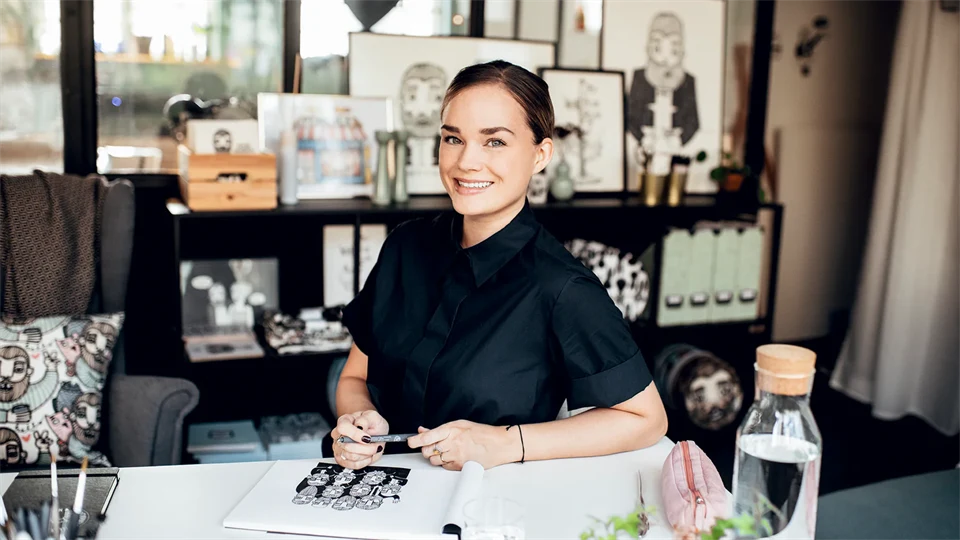 A woman in a black shirt is sitting in a room with a bookshelf behind her.