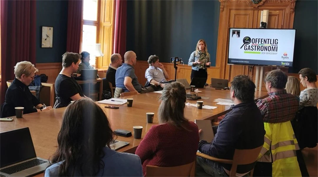 Two dozen people in a room with a woman at the front presenting in front of a screen
