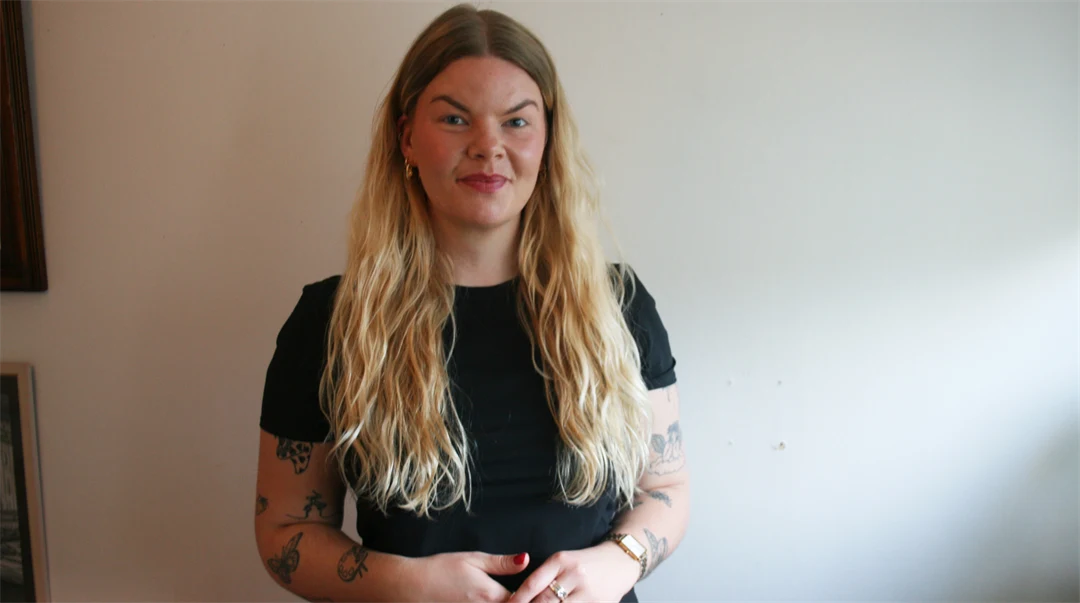 Kvinna i svart kortärmad tröja och långt blont hår står framför en beige vägg. 