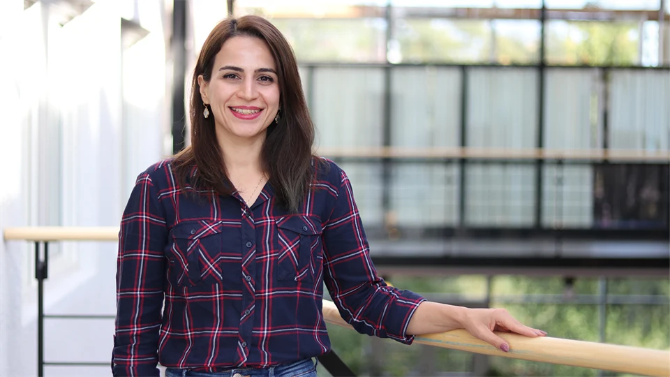 A woman in a checkered blouse stands by a railing and smiles at the camera.