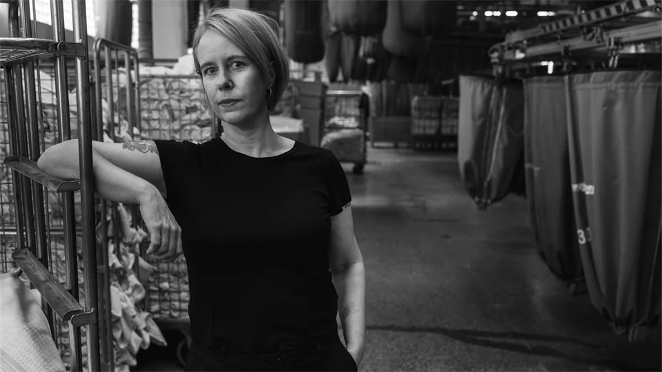 En kvinna med ljust hår, klädd i svart, poserar framför kameran, i bakgrunden syns vagnar med tvätt.