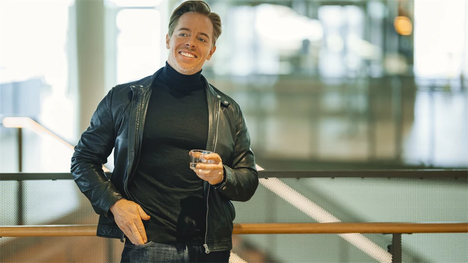 A smiling man in a black turtleneck and a black leather jacket is leaning against a railing. He is holding a glass of water. 