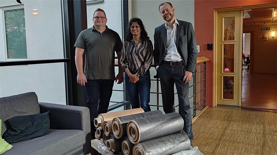  Three people stand together behind a stack of silver-grey large rolls of paper.