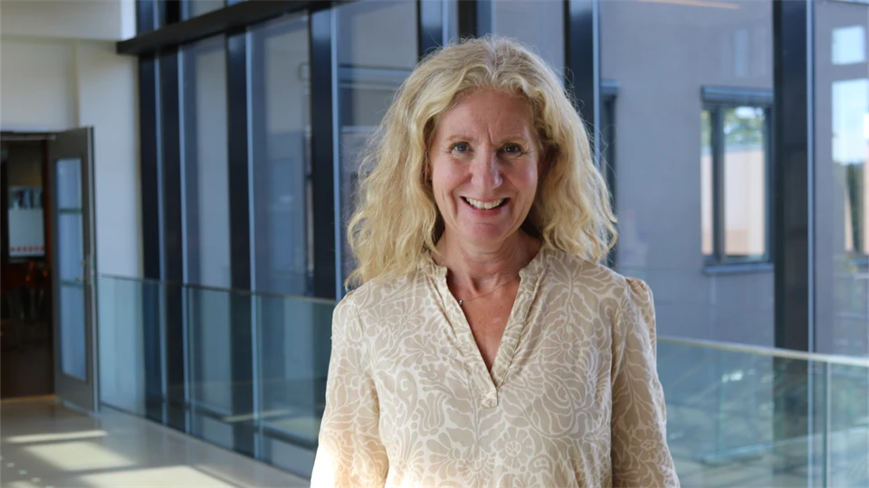 En kvinna med ljust hår och cremevit blus står i en glaskorridor med fönster bakom sig.