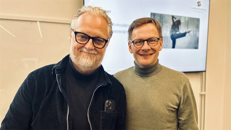 Två män i glasögon tittar in i kameran och ler. De står framför en skärm. 