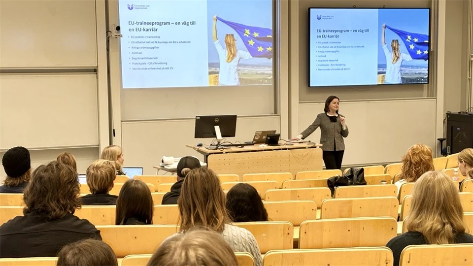 En kvinna i mörkt kort hår står i en föreläsningssal, studenter sitter på stolar vänd mot henne