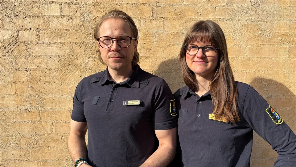 Två personer står framför en beige husfasad iklädda uniformer från Kriminalvården