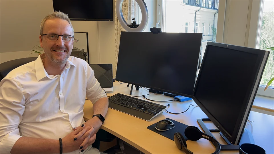  A man in glasses and wearing a white shirt sits in front of some computer screens.