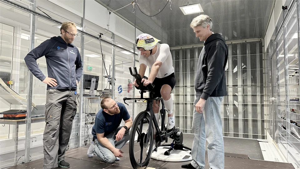 Tre män står och tittar på en fjärde man som sitter på en cykel inomhus.