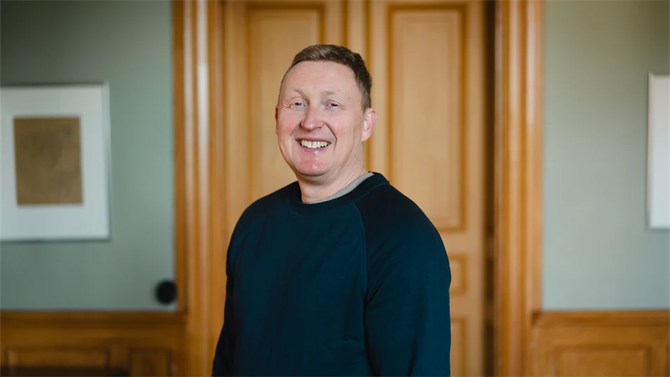 Man i blå collegetröja står framför en brun trädörr och ler mot kameran.