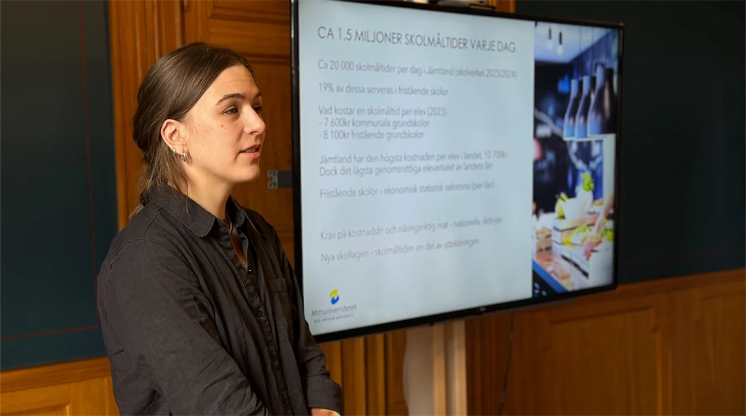 En kvinna i brunt hår och mörkgrå skjorta står vid en skärm med en presentation på. 