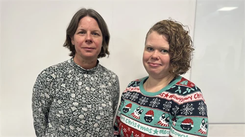 Två kvinnor, en i blommig tröja och med kort, brunt hår och en kvinna med blont, axellångt hår i stickad tröja med julmotiv.