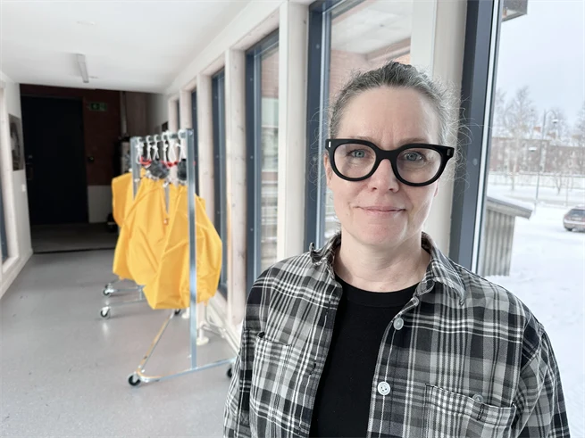 A woman dressed in a checkered shirt and black frames stand in a corridor with windows.