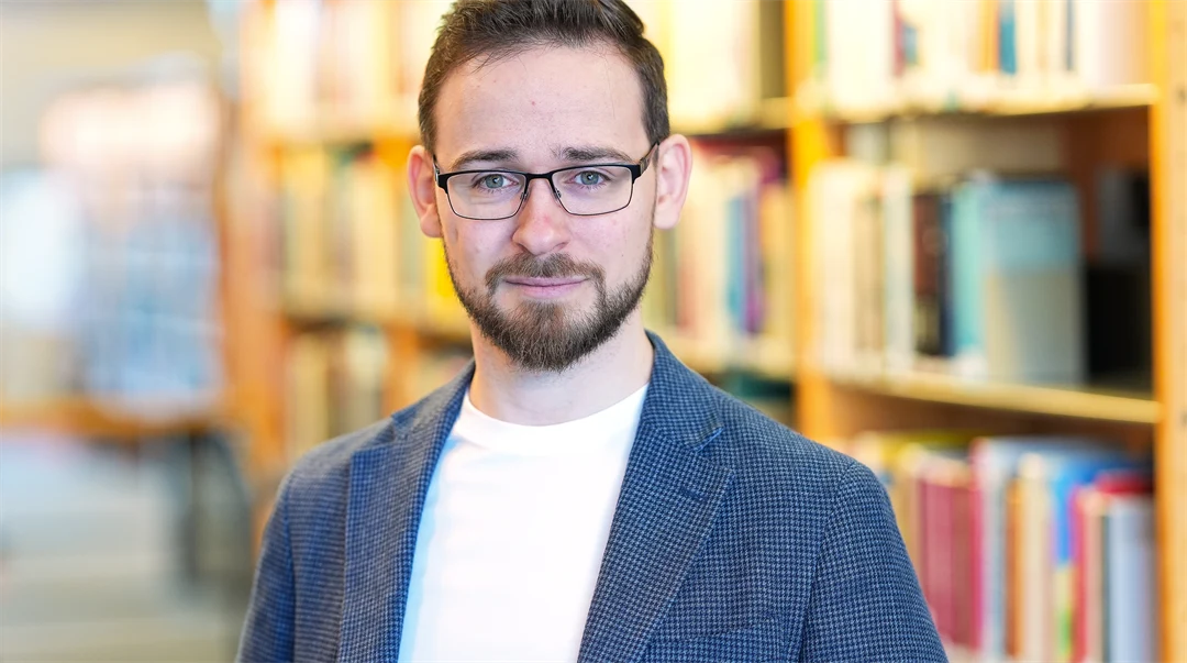 Porträtt av en man i mörkt hår, skäggstubb, glasögon. Klädd i vit t-shirt och blå kavaj