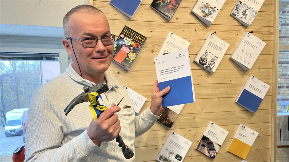 En man har en avhandling i ena handen och en hammare i den andra handen. Han står framför en vägg med uppspikade avhandlingar.